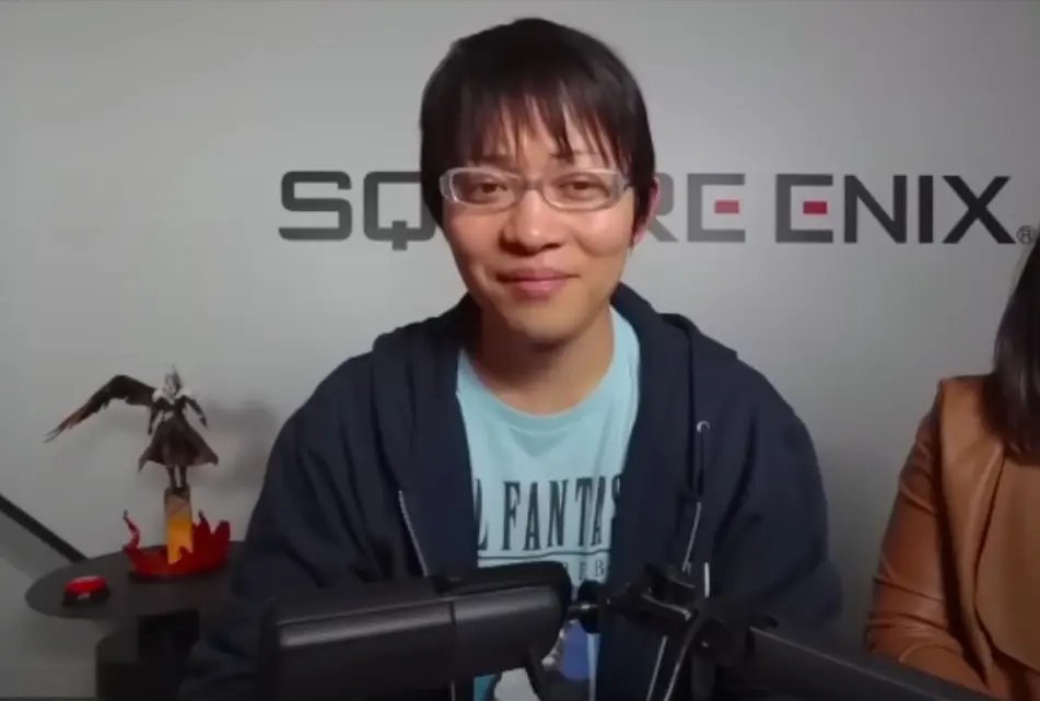 Naoki Hamaguchi portant des lunettes et un t-shirt bleu, assis devant un mur Square Enix.