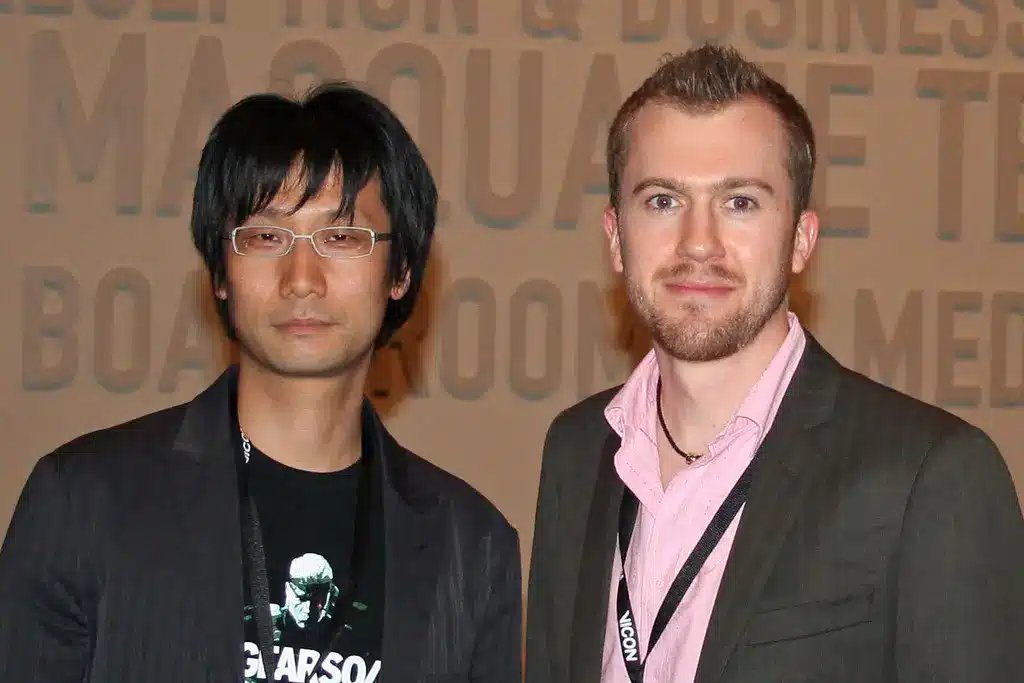 Hideo Kojima portant des lunettes et un blazer noir, debout à côté d'un autre homme.