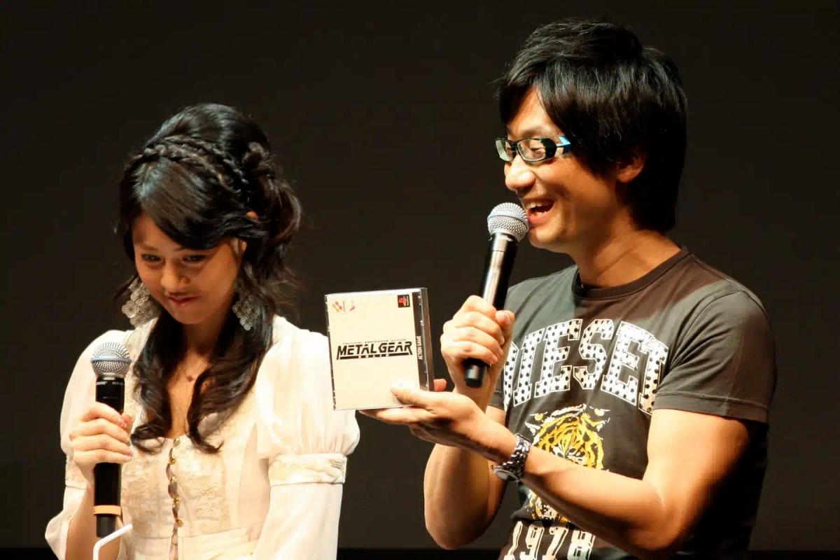 Hideo Kojima tenant une boîte de jeu Metal Gear tout en parlant dans un micro.